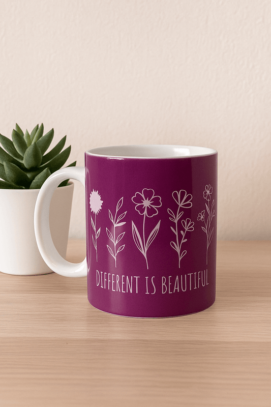Purple mug with floral design and 'Different is Beautiful' text on a wooden surface with a plant in the background.